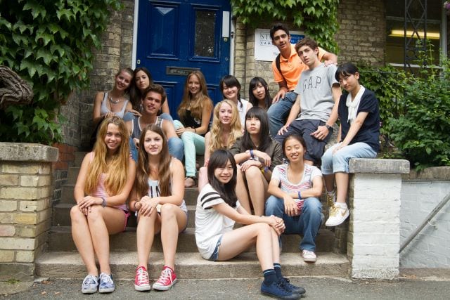 Large group of students chat and laugh on steps outside Studio Cambridge Main School, where English lessons for Sir Michael English summer camp are held