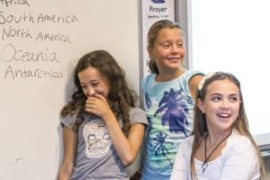 Three students laugh by the whiteboard in English language class at Sir Henry residential English summer camp. Visible writing on the board reads: 'South America, North America, Oceania, Antarctica'.