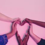 Six hands of people with a variety of different skin tones coming together in the shape of a heart on a dusty pink background.