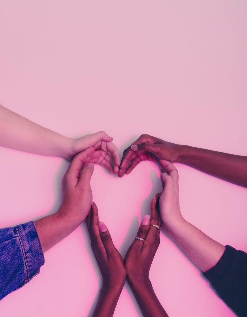 Six hands of people with a variety of different skin tones coming together in the shape of a heart on a dusty pink background.