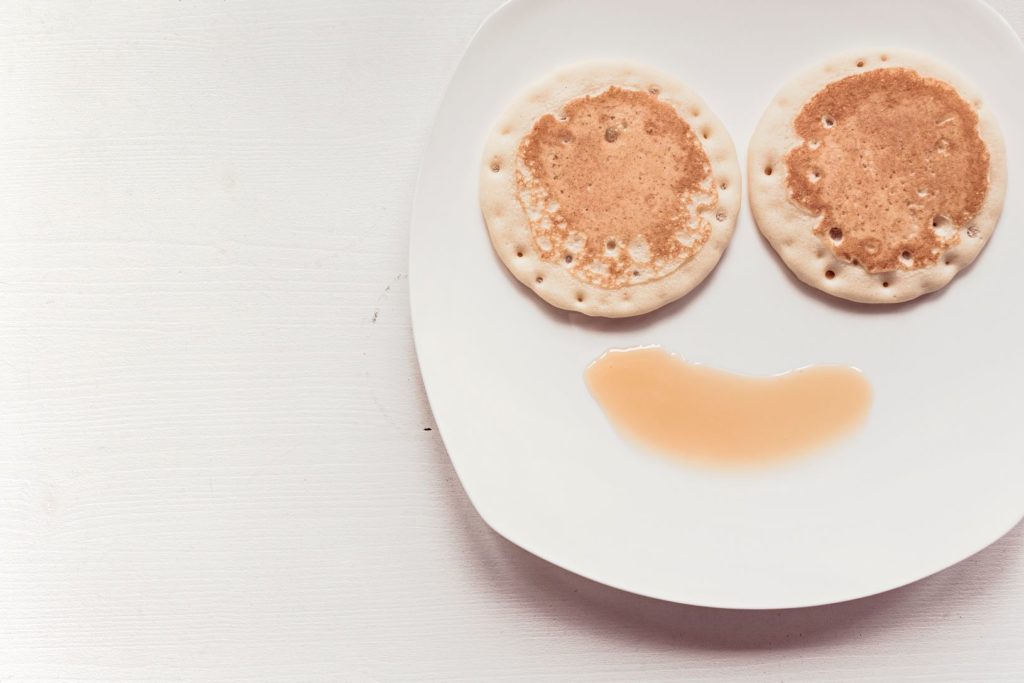 Two pancakes on a white plate with a splash of syrup underneath, arranged to look like a smiley face.