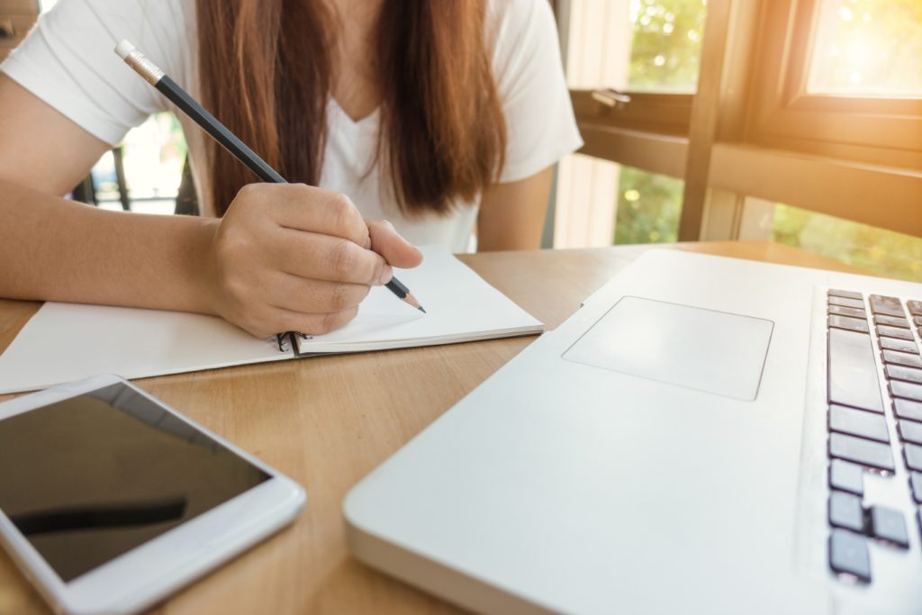 English student sits at a desk taking notes from their laptop with their phone beside them